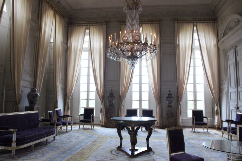 Room with chandelier and tall yellow curtains with the sun streaming in the room at Versailles