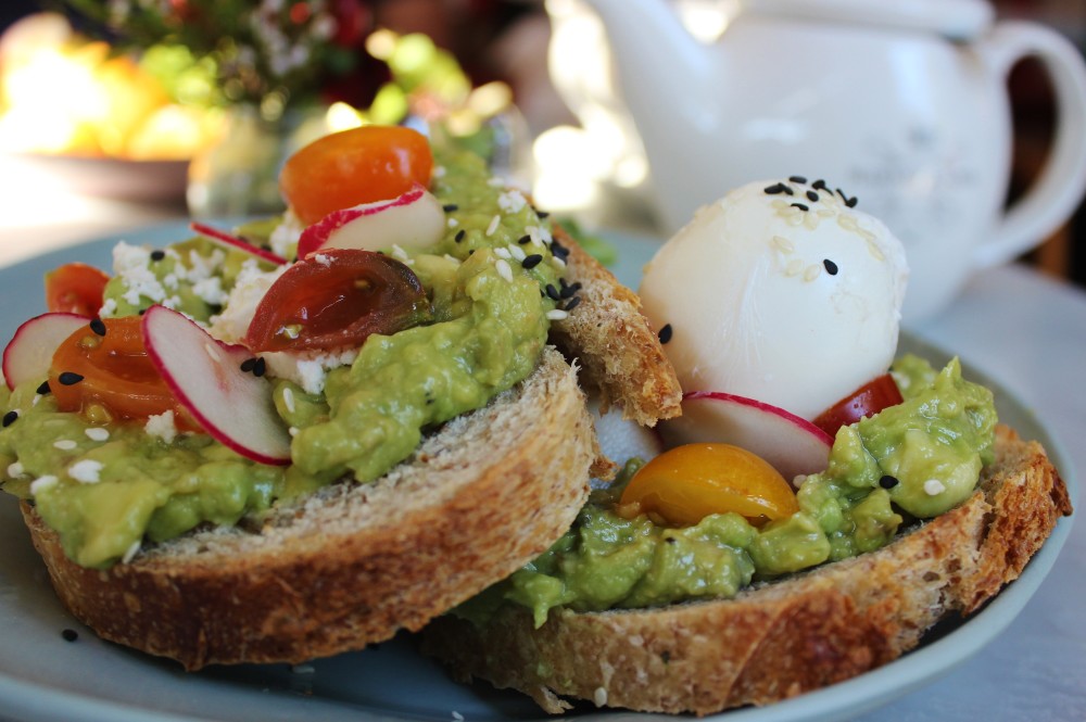 avocado toast with cherry tomatoes, radish slices, and a poached egg. A teapot sits in the background.